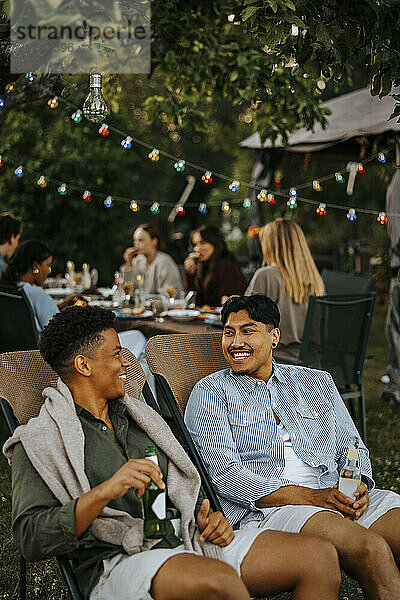 Happy male friend enjoying drinks while sitting on chair and talking in back yard at social party