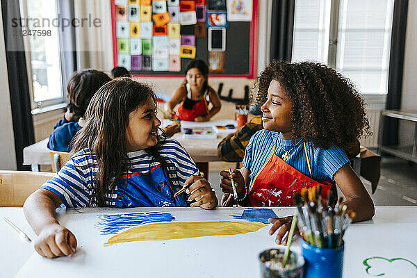 Smiling curly hair girl with female classmate sitting in art class at elementary school