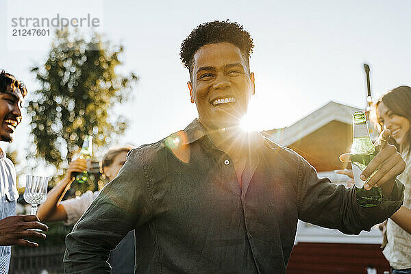 Portrait of happy young man holding beer bottle while dancing in back yard on sunny day at social party