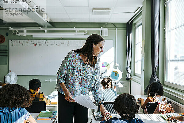 Smiling female professor distributing test papers to pupils sitting in classroom at school