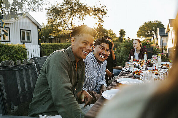 Happy male friends talking while sitting near dining table in back yard at dinner party