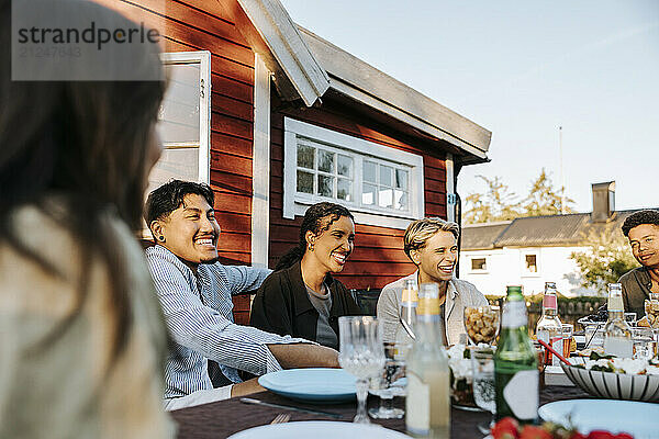 Happy male and female friends sitting together at dining table in back yard for dinner party