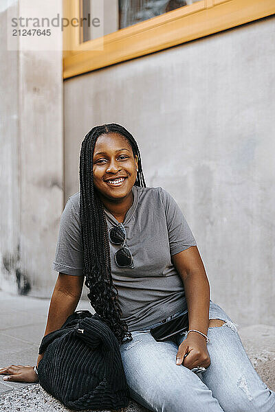 Portrait of smiling braided hair girl sitting with bag near wall of school building