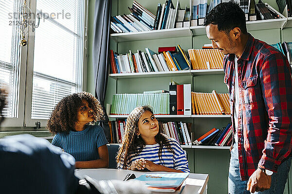 Side view of male professor assisting elementary schoolgirls sitting in library