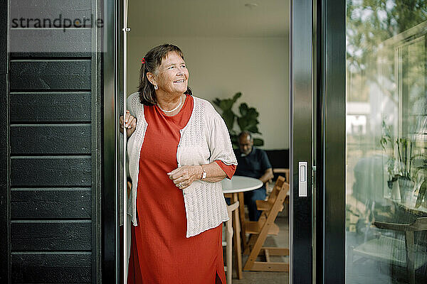 Smiling senior woman looking away while standing at house doorway