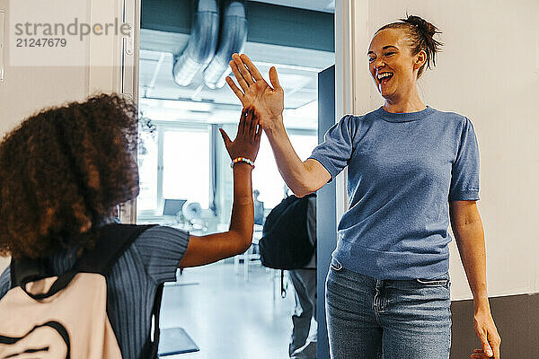 Cheerful female teacher giving high-five to female pupil while standing near doorway of classroom in school