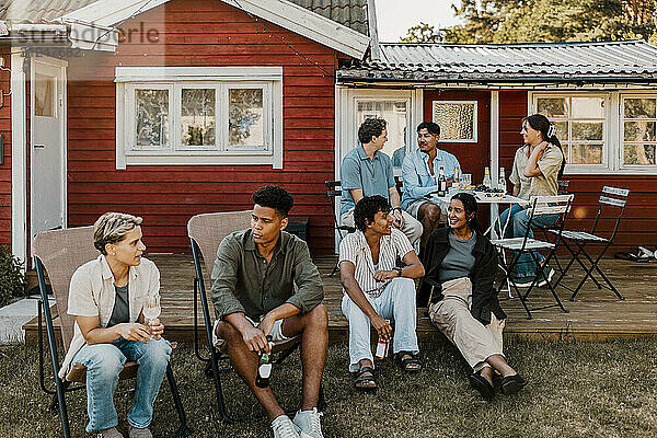 Multiracial group of male and female friends sitting and talking with each other while sitting in back yard at social pa