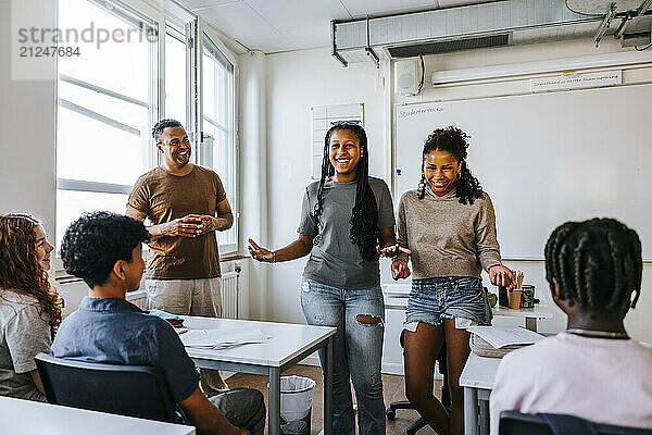 Happy teenage girls standing while talking with students in classroom at junior high school