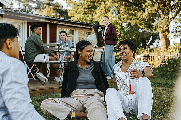 Happy male and female friends having drinks while sitting on grass in back yard at dinner party