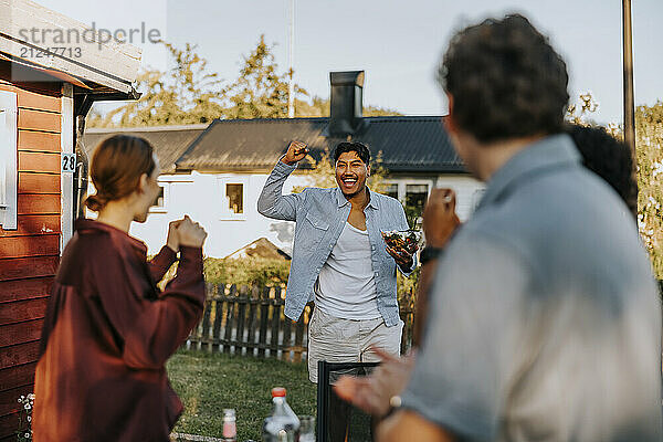 Cheerful young man holding fist and desert bowl while walking towards friends in back yard at dinner party