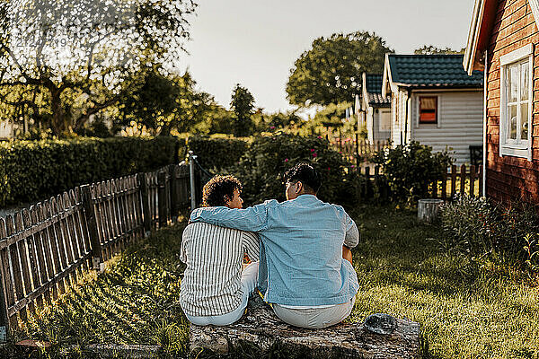 Rear view of young man with arm around male friend sitting on log in back yard at sunny day