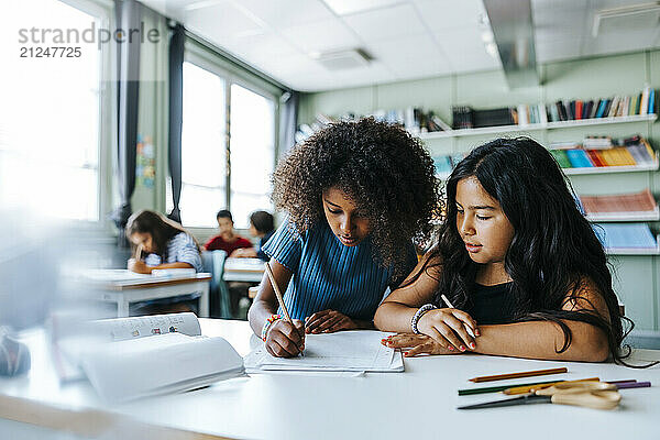 Curly hair girl writing while sitting with female classmate near desk in classroom at elementary school