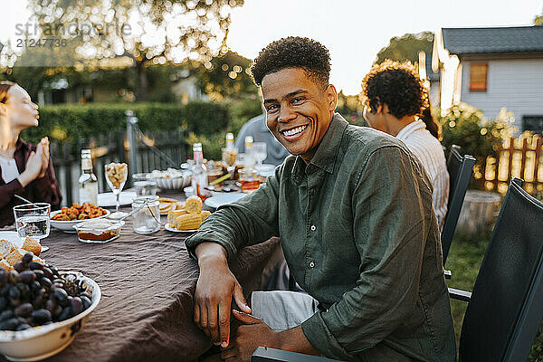 Portrait of happy curly hair young man sitting near dining table in back yard at dinner party