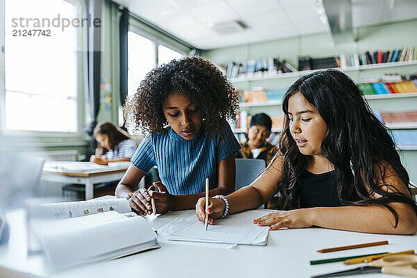 Female classmates writing while studying together and sitting near desk in classroom at elementary school