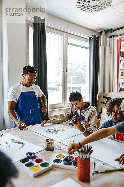 Smiling male teacher assisting pupil painting while sitting in art class at elementary school