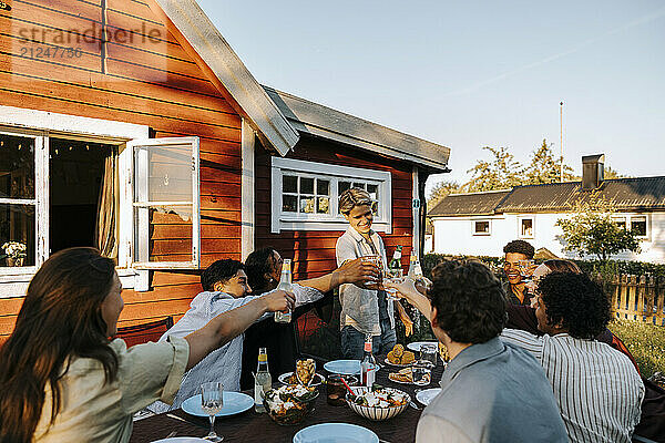 Happy young woman doing celebratory toast with male and female friends sitting near dining table in back yard