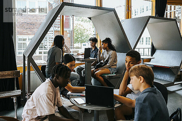 Group of male and female students studying together through laptop while sitting in cafeteria at school