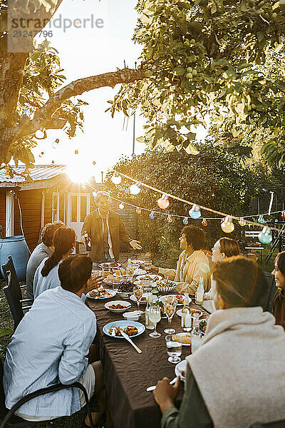 String light decoration with young man giving speech to male and female friends at dinner party in back yard