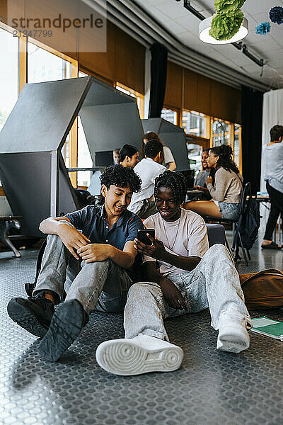 Happy male friends using smart phone while sitting in cafeteria at school