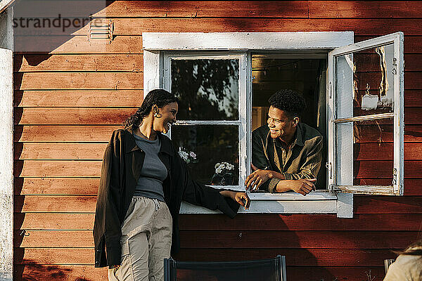 Smiling young man leaning on window and talking with female friend standing in back yard