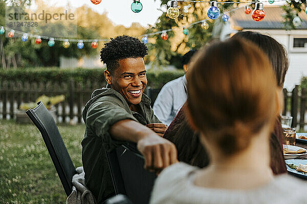 Happy young man sitting on chair while talking with female friend in back yard at dinner party