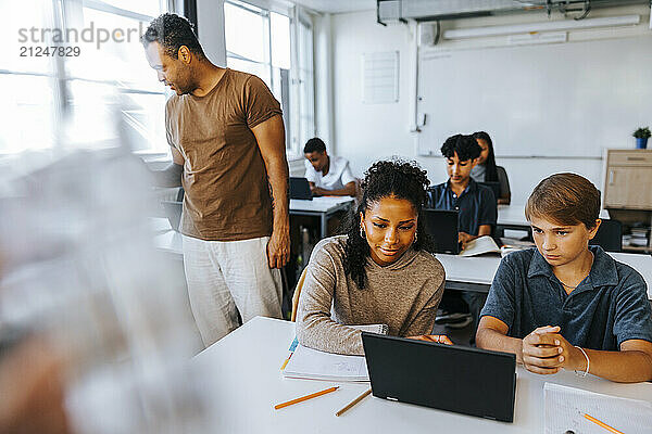 Teacher standing near male and female students doing e-learning in classroom at junior high school