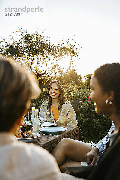 Portrait of smiling young woman sitting near dining table with friends in back yard at dinner party