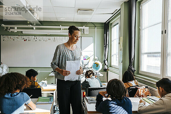 Female teacher holding test papers while giving instructions to group of students sitting in classroom at school