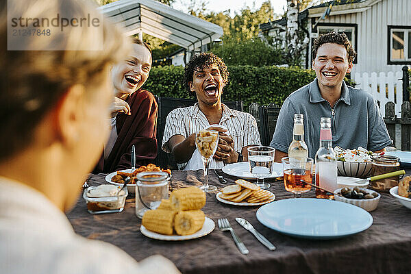 Cheerful male and female friends having dinner while sitting near table in back yard at social party