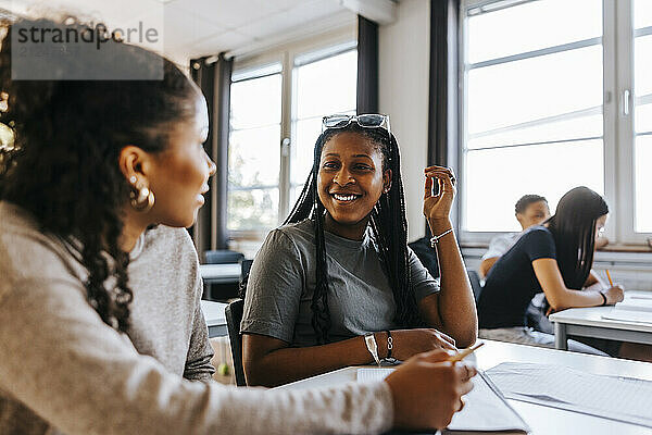 Happy teenage girl with braided hair talking with female friend while sitting in classroom at junior high school