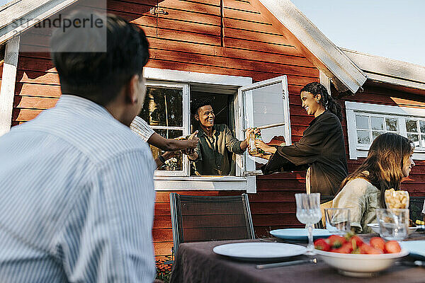 Smiling young man passing beer bottles from window to friends standing in back yard at dinner party