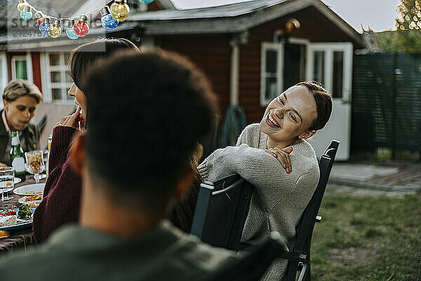 Happy young woman leaning on chair while talking with male friend in back yard at dinner party