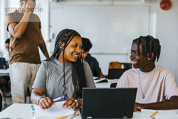 Happy male and female junior high students studying together while sitting with laptop near desk in classroom at school