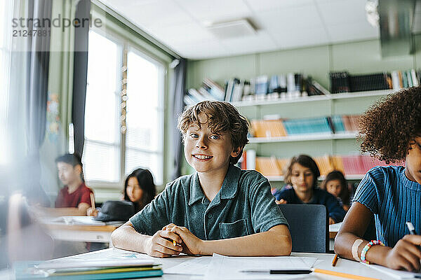 Portrait of cute boy sitting in classroom with classmates at elementary school