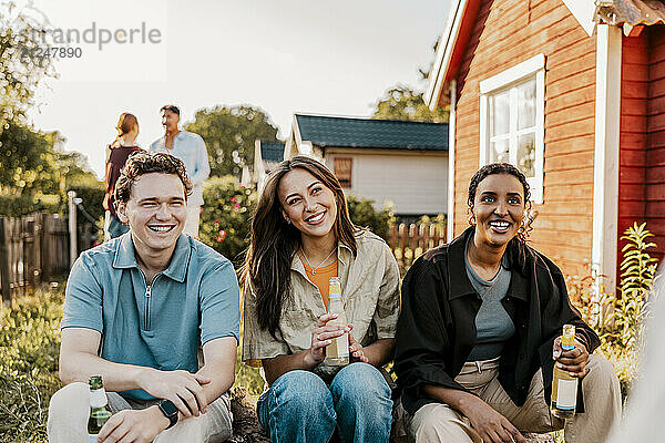 Happy male and female friends holding drinks while sitting in back yard at social gathering