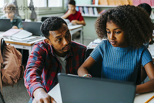 Male professor helping curly hair student with studies on laptop in classroom at elementary school