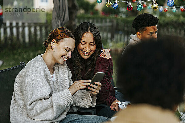 Female friends using smart phone while sitting on chair in back yard at social party