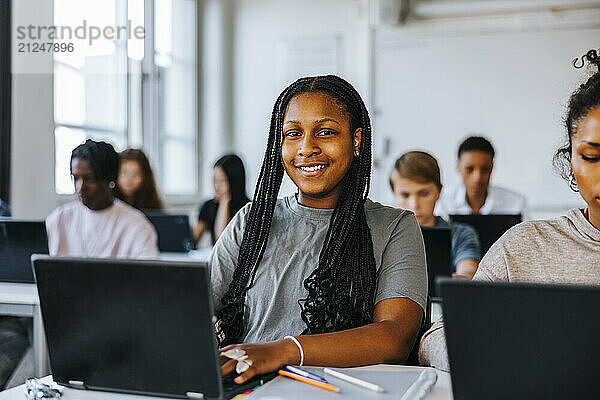 Portrait of teenage student with braided hair sitting with laptop in classroom at junior high school