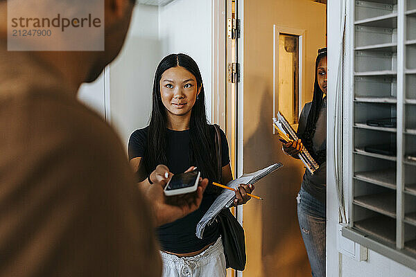 Professor collecting smart phone from female student standing near doorway of classroom in school