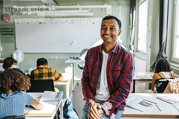 Portrait of happy male teacher in plaid shirt sitting on desk in classroom at elementary school