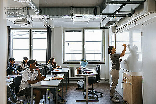 Female professor writing on whiteboard while teaching junior high students in classroom at school