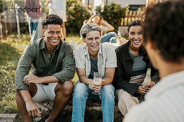 Happy male and female friends holding drinks while sitting on log in back yard at social gathering