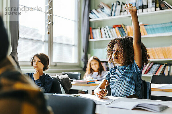 Curly hair girl with hand raised sitting in classroom at school