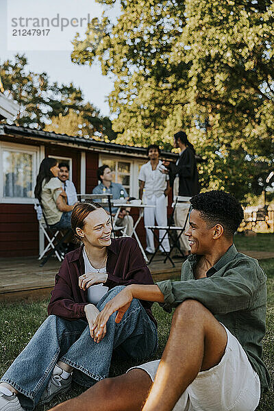 Smiling young woman talking with male friend while sitting on ground in back yard at social gathering