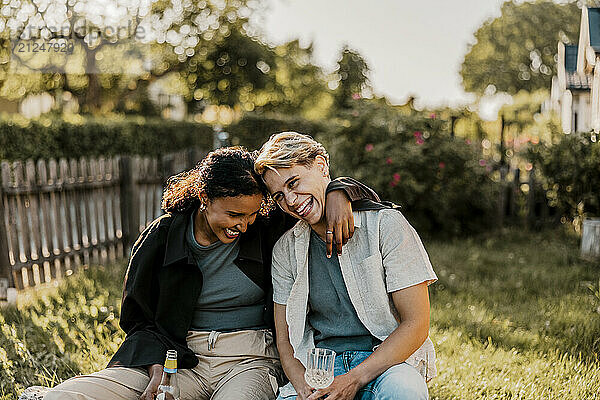 Carefree young woman with arm around female friend enjoying drinks in back yard at sunny day