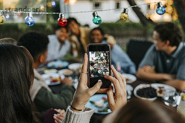 Woman photographing male and female friends through smart phone in back yard at dinner party