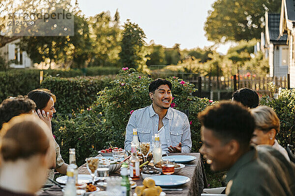 Smiling young man talking with friend while sitting near dining table in back yard at dinner party