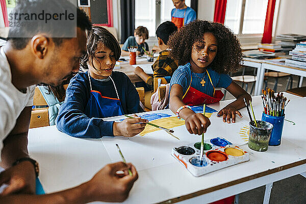 Art professor leaning on desk and teaching pupils painting while sitting in classroom at school