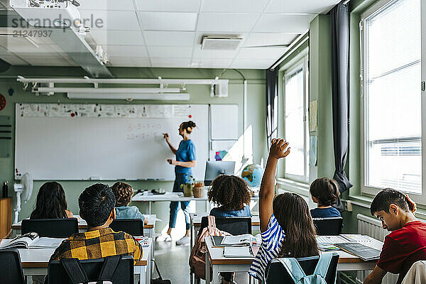 Rear view of girl with hand raised sitting in classroom while female teacher teaching at school