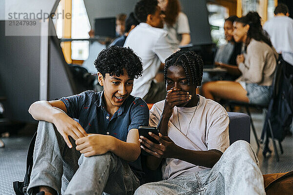 Shocked male friends using smart phone while sitting in cafeteria at school